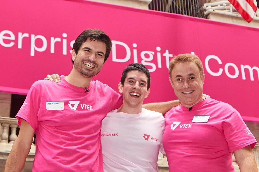 Guilherme Rodrigues between Geraldo Thomaz and Mariano Gomide, in VTEX t-shirts, in front of VTEX's pink banner at the NYSE.